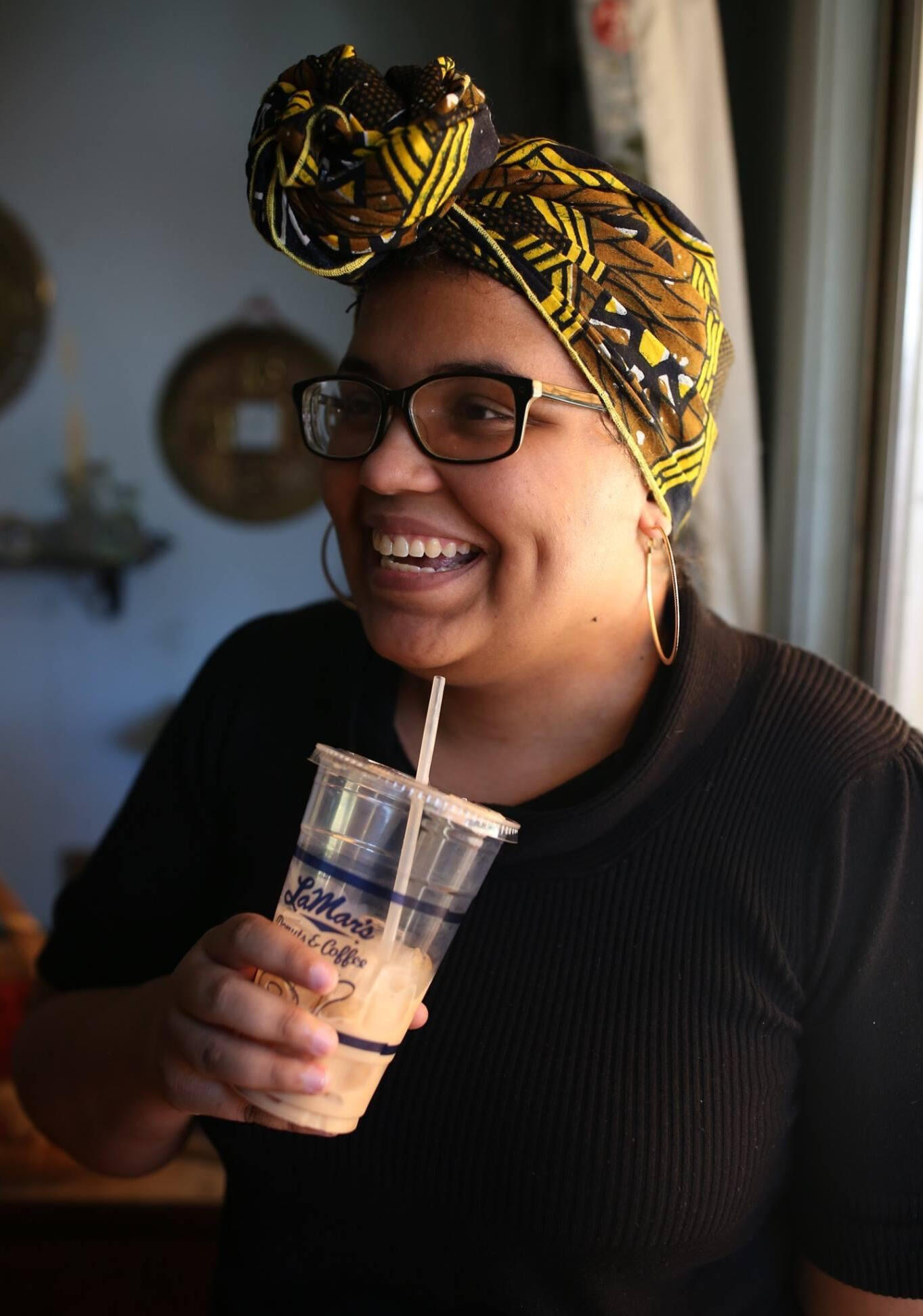 A color photo of Bunny, a light-skinned Black femme person. She is wearing black square-ish glasses, a black knit dress, simple gold hoop earrings, and a black, brown, and yellow headwrap with a knot at the front. She is smiling and looking to the left off