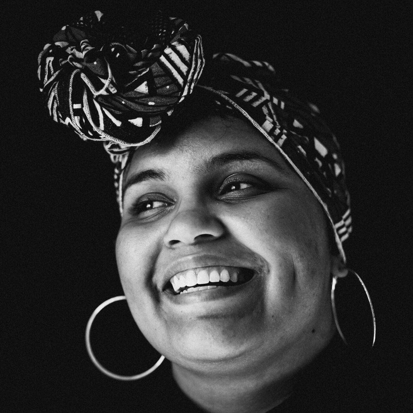 A black and white portrait of Bunny wearing a patterned head wrap with a front knot, gold hoop earrings, smiling brightly as she looks to the left.